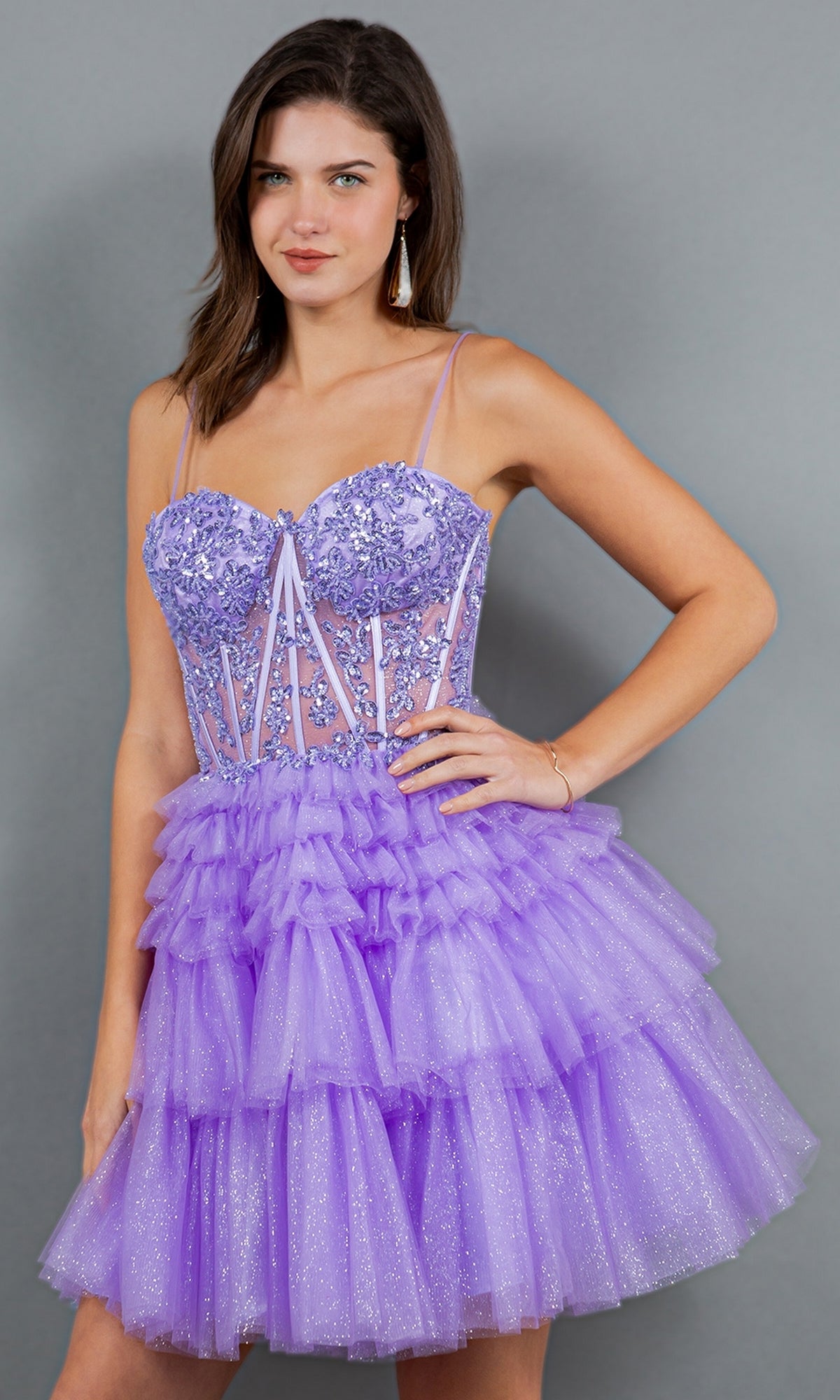 A woman models the Short Sweetheart Ruffled Homecoming Dress 5133J, featuring spaghetti straps, floral bodice embellishments, and a tiered ruffled skirt in lavender, set against a gray background.